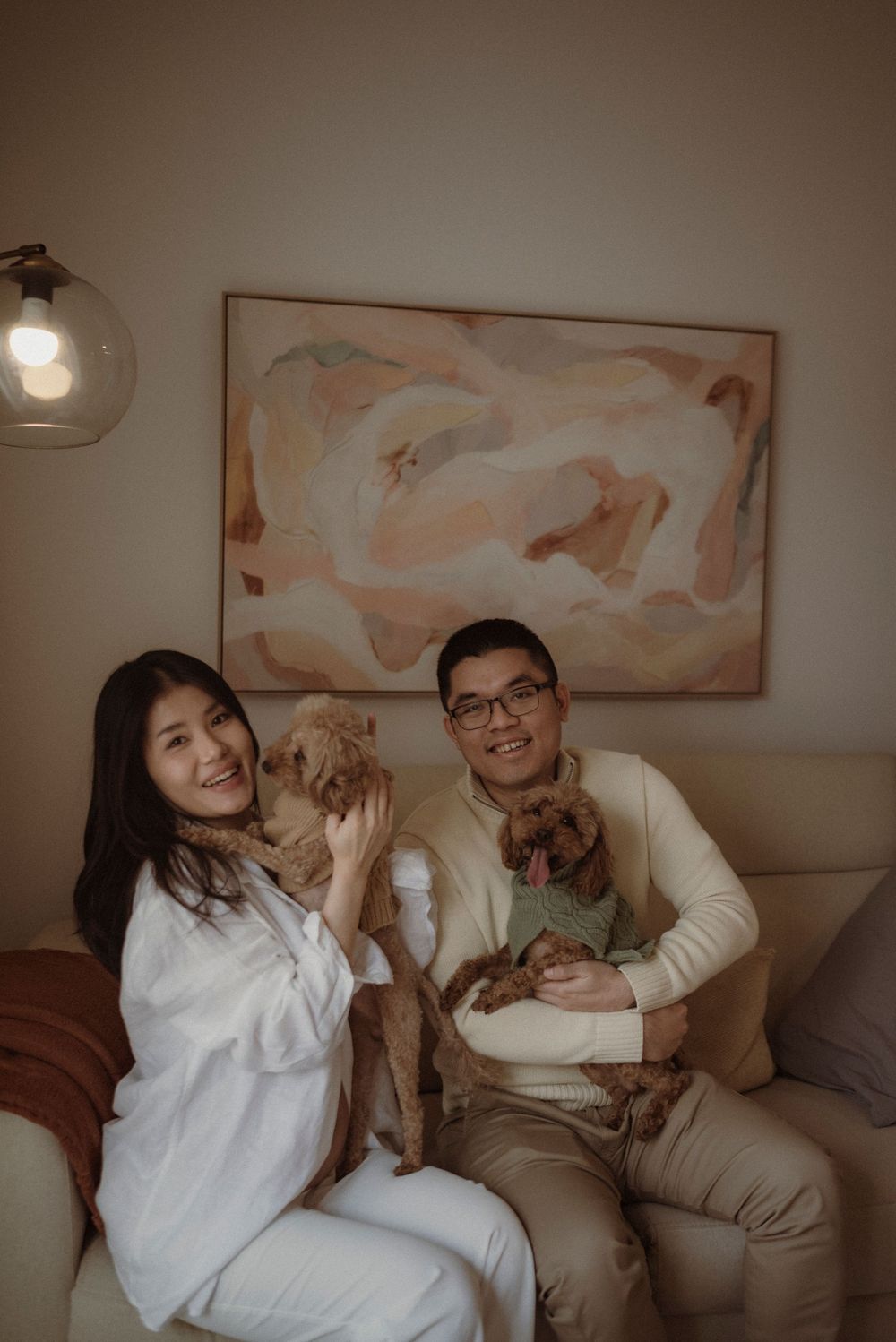 A smiling couple sits on a beige sofa holding two brown dogs, with abstract pastel artwork on the wall behind them.