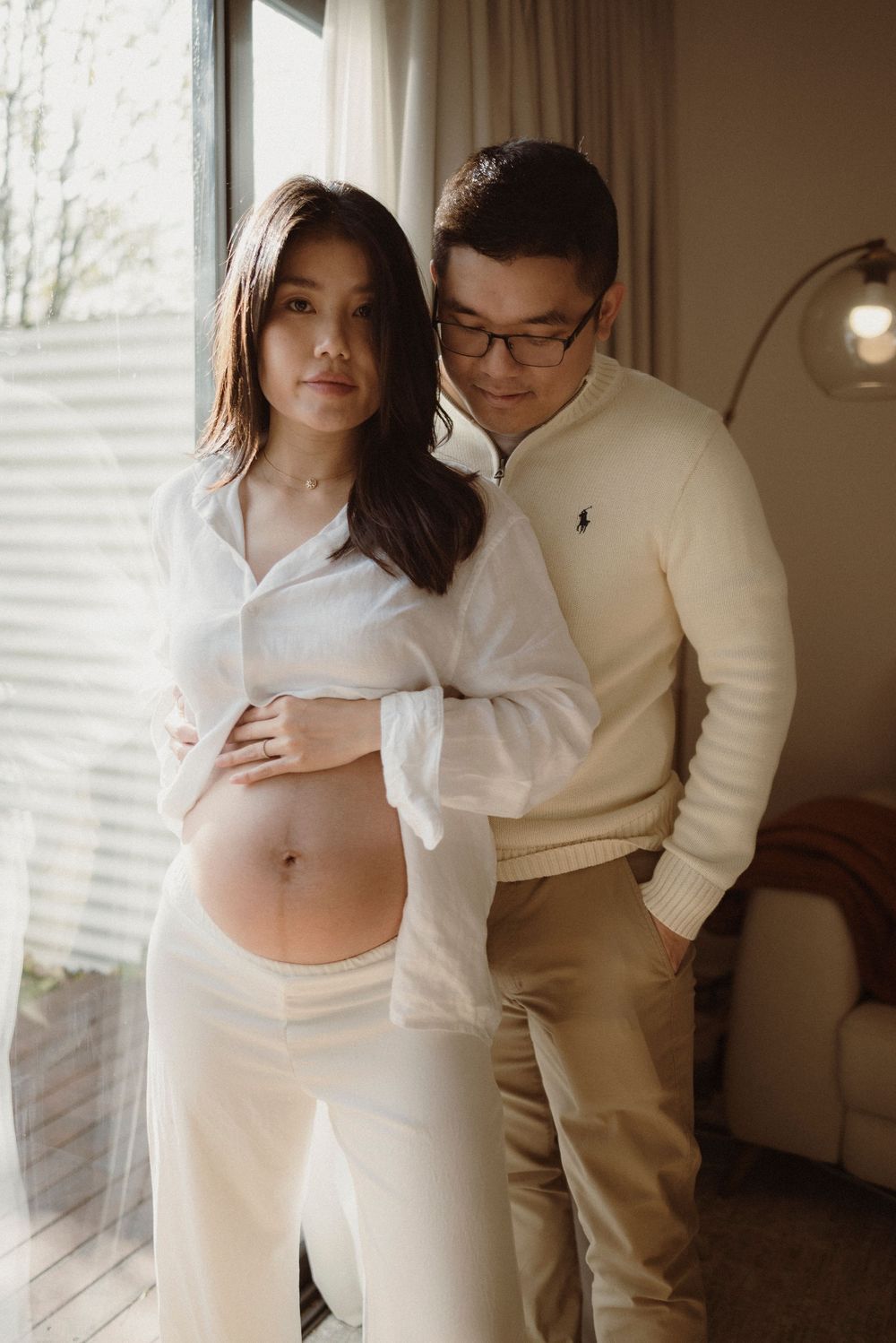 A pregnant woman in a white shirt poses with her partner in a cozy indoor maternity photoshoot.
