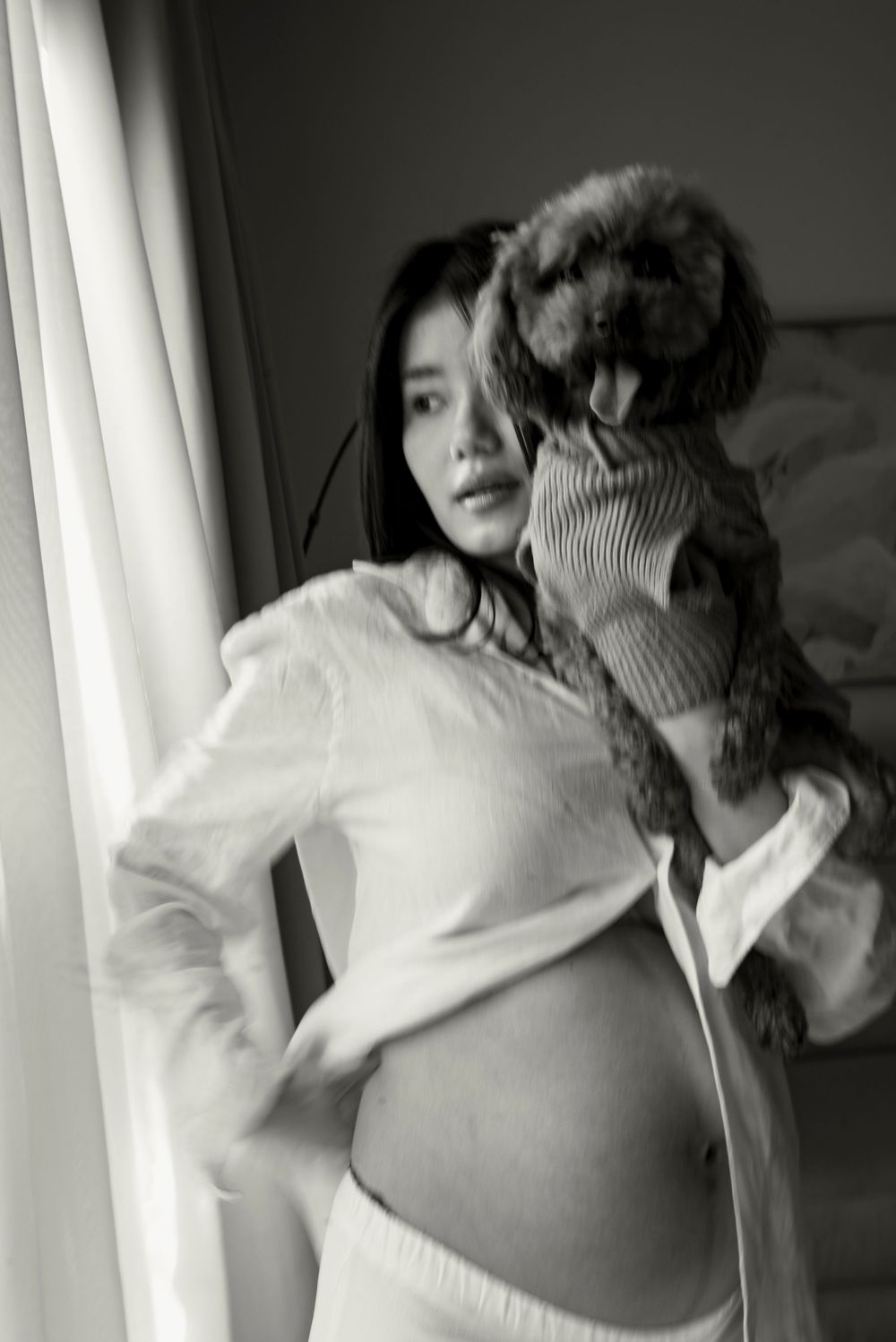 Black and white photo of a pregnant woman holding a small dog near a window, wearing a white top.