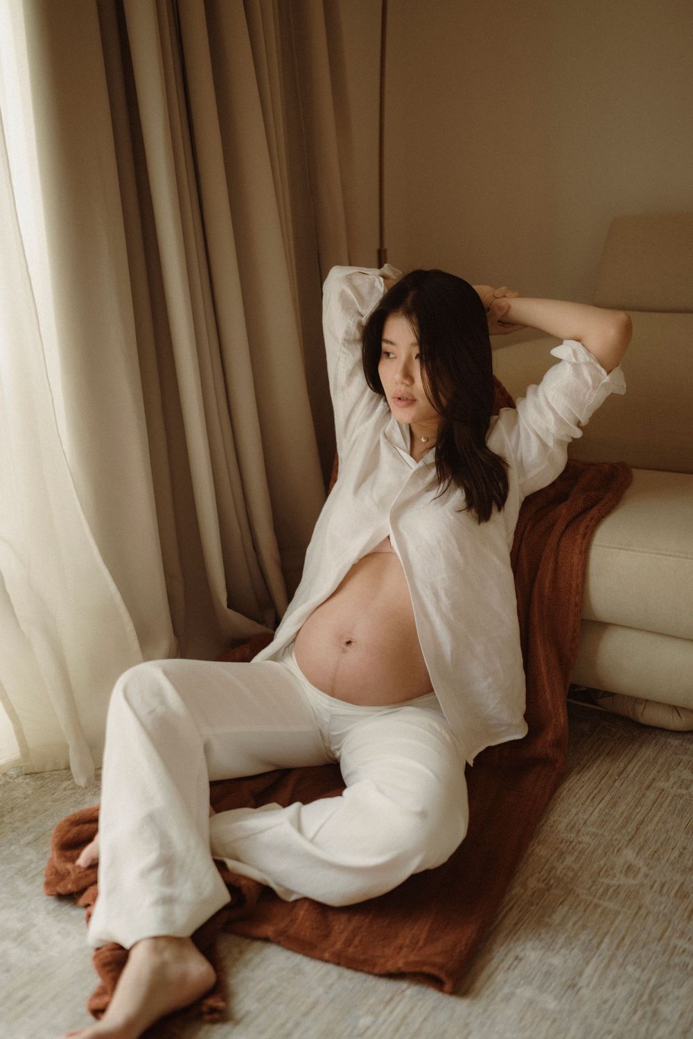 Pregnant woman in white linen outfit sitting on floor, posing for maternity photoshoot in neutral toned room.