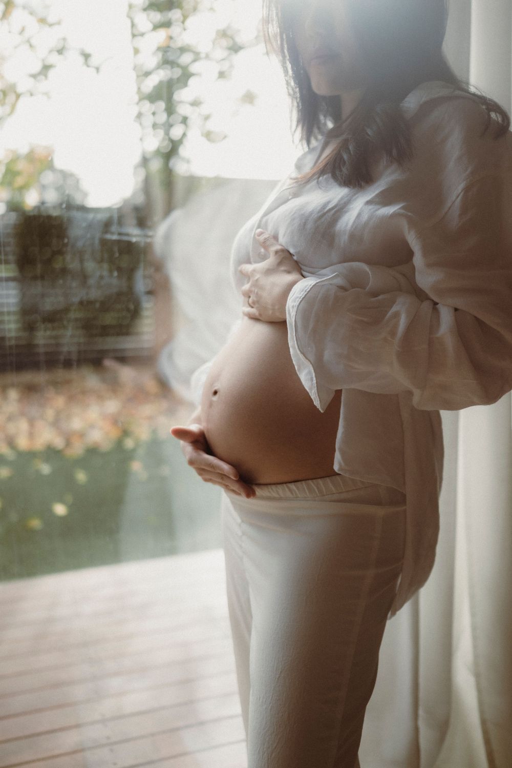Pregnant woman in white shirt cradling bare baby bump near sunlit window with autumn trees outside.