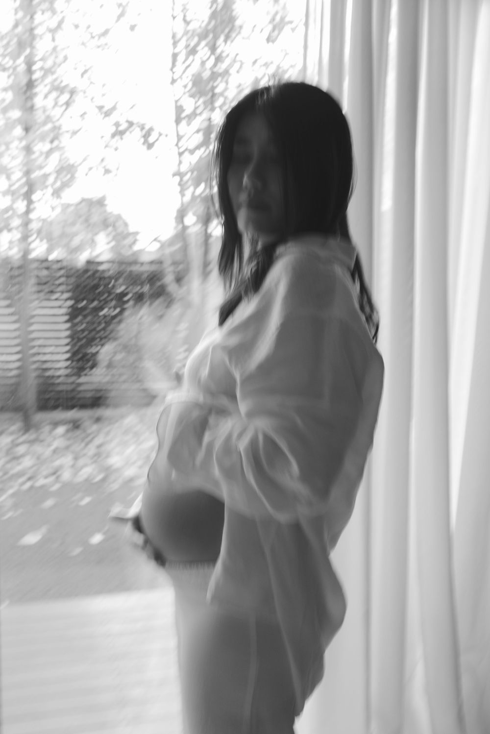 Black and white photo of a pregnant woman in a sheer white shirt standing by a window with curtains.