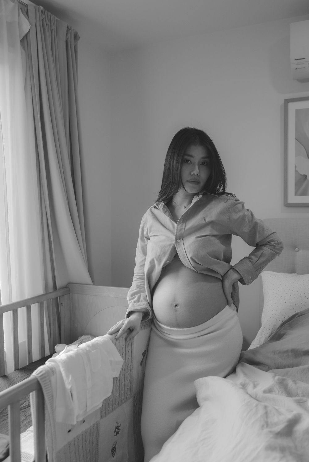 Black and white photo of a pregnant woman posing in a bedroom, wearing an open shirt and skirt, showing her baby bump.