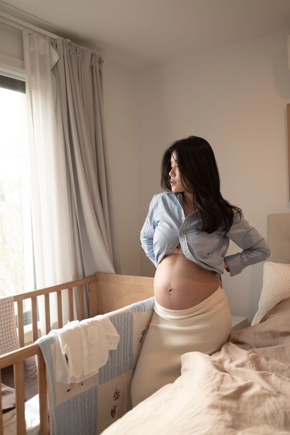 Pregnant woman in open denim shirt standing beside wooden crib in bright bedroom, showing baby bump.