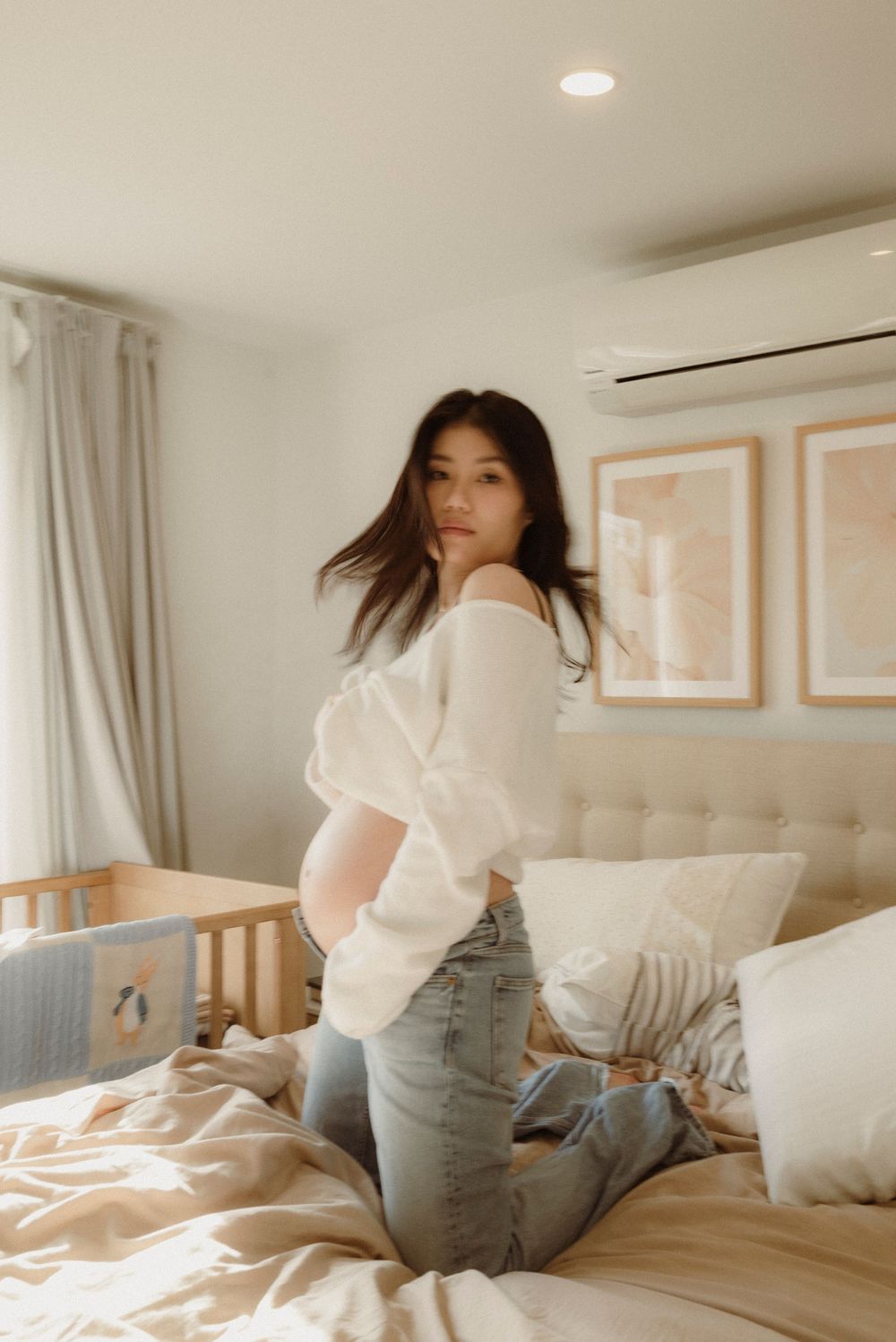 Pregnant woman in white sweater and jeans kneeling on bed, showing baby bump in bright modern bedroom.