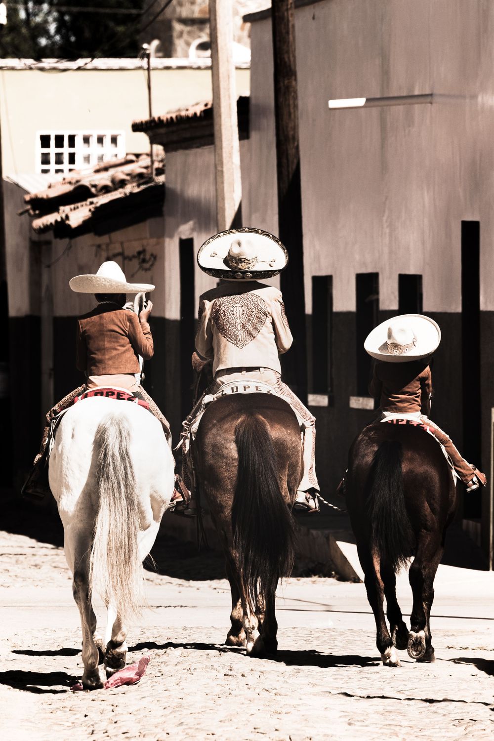 Three charros in traditional Mexican attire and sombreros ride horses down a sunlit cobblestone street.