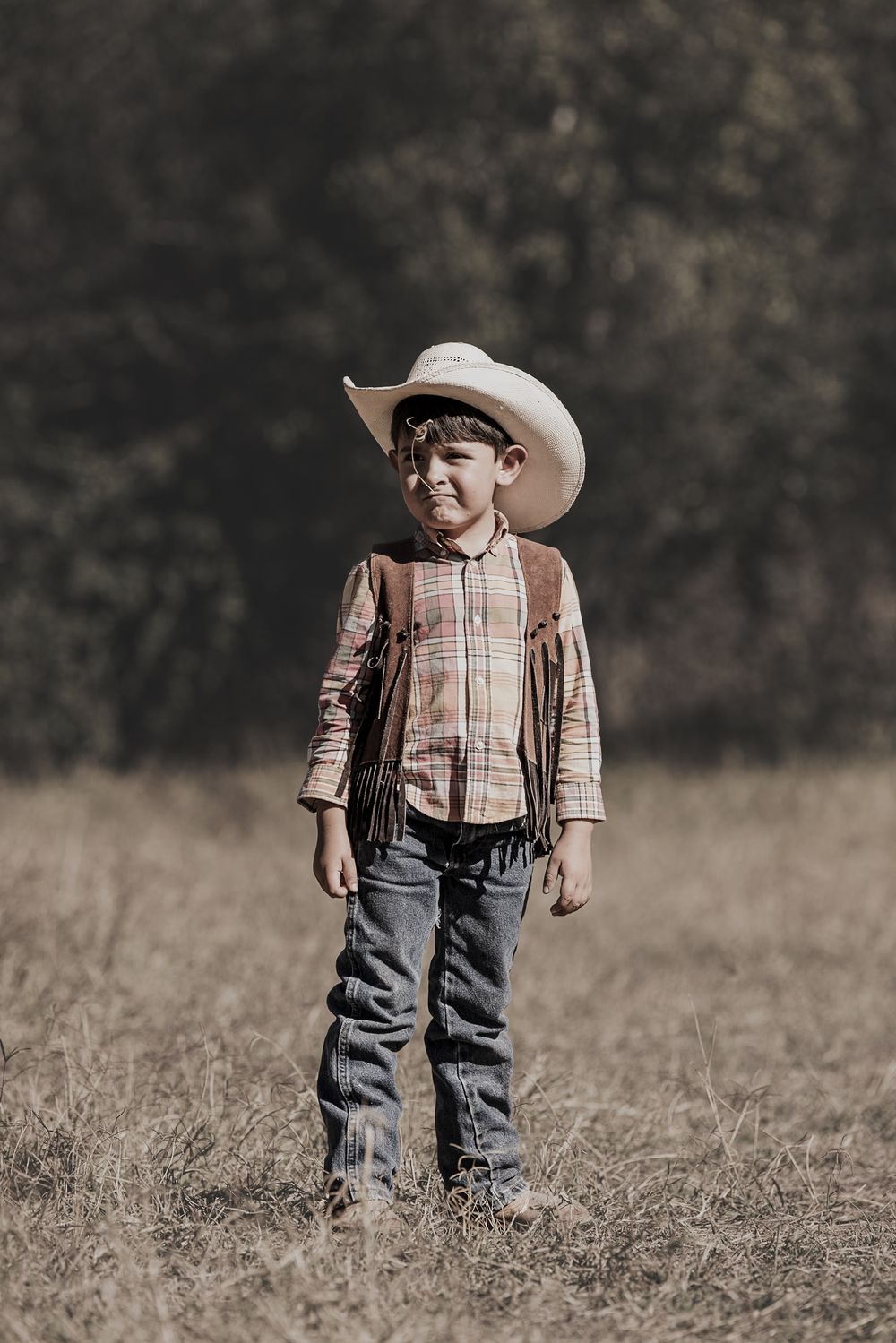 Young boy in oversized cowboy hat and plaid shirt with leather vest standing in field.