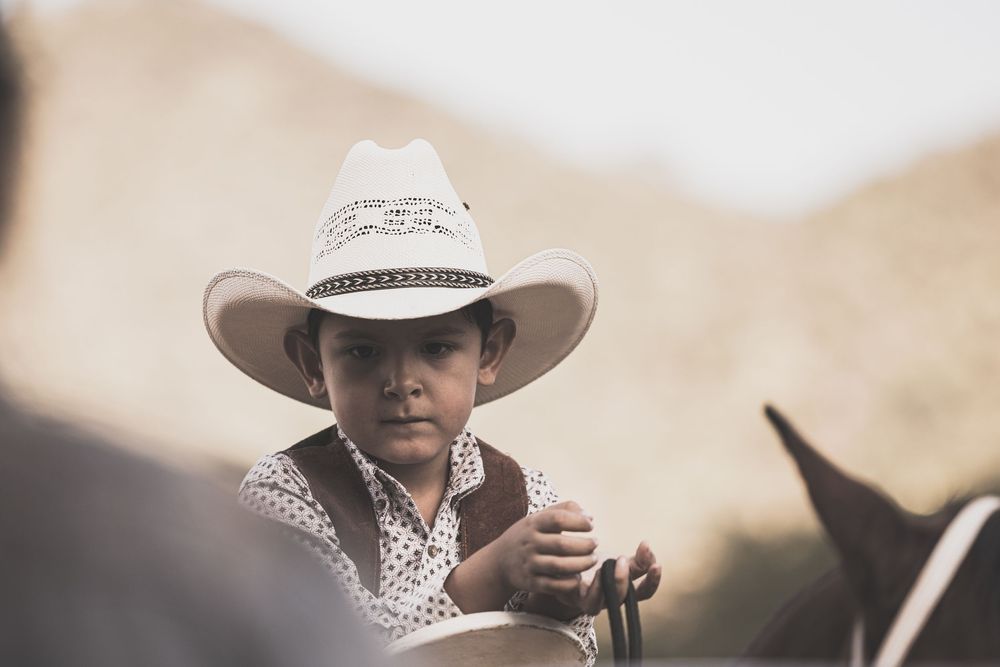 Young child wearing a white cowboy hat sitting on a horse at a western rodeo event in a desert setting.