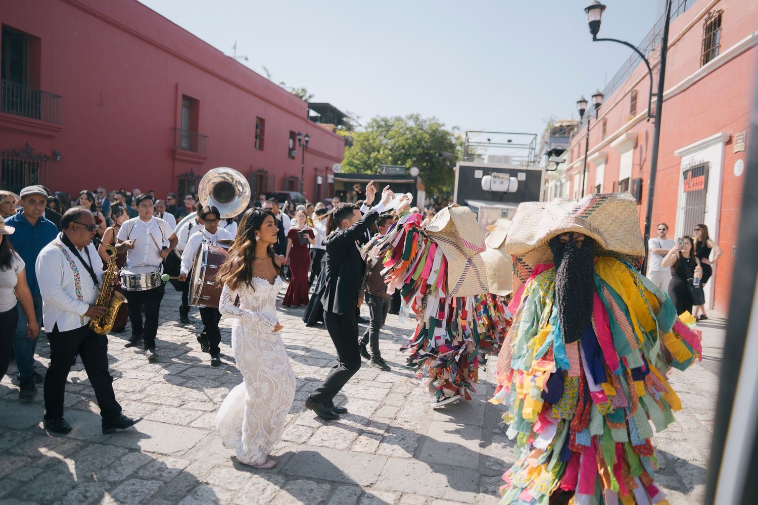 Colorful Mexican wedding procession with bride and groom dancing through cobblestone streets with a brass band and festive costumes.