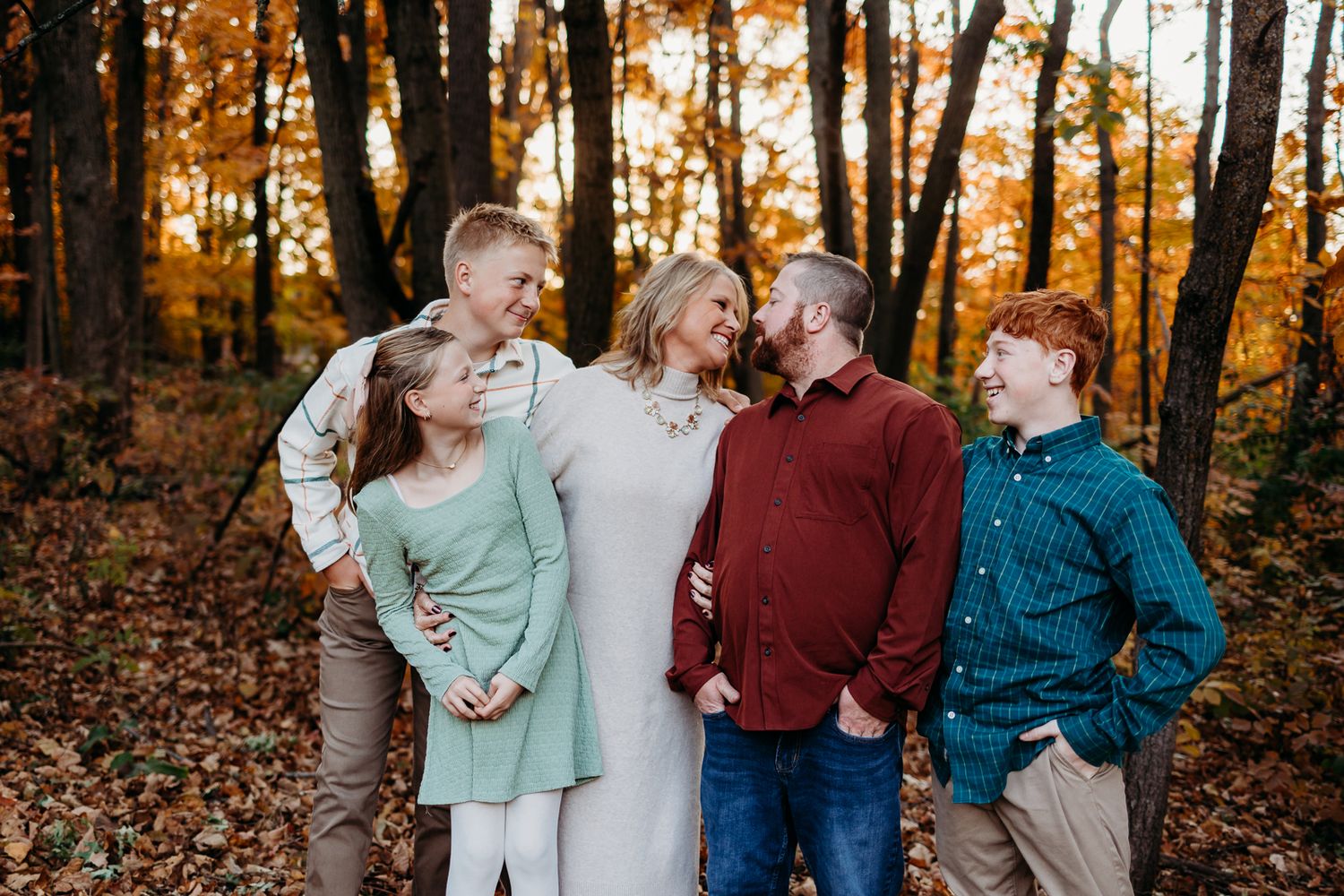 A family of 5 looking at each other laughing and smiling. There are leaves on the ground and they are standing in a forest.