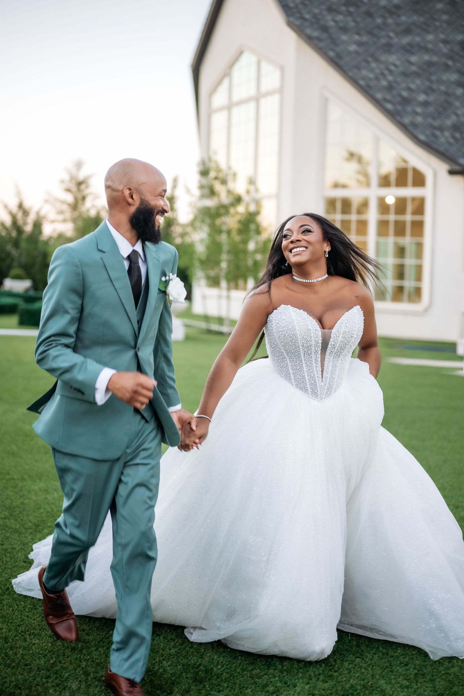 A joyful bride in a white ballgown and groom in a sage suit hold hands walking outdoors at their wedding venue.