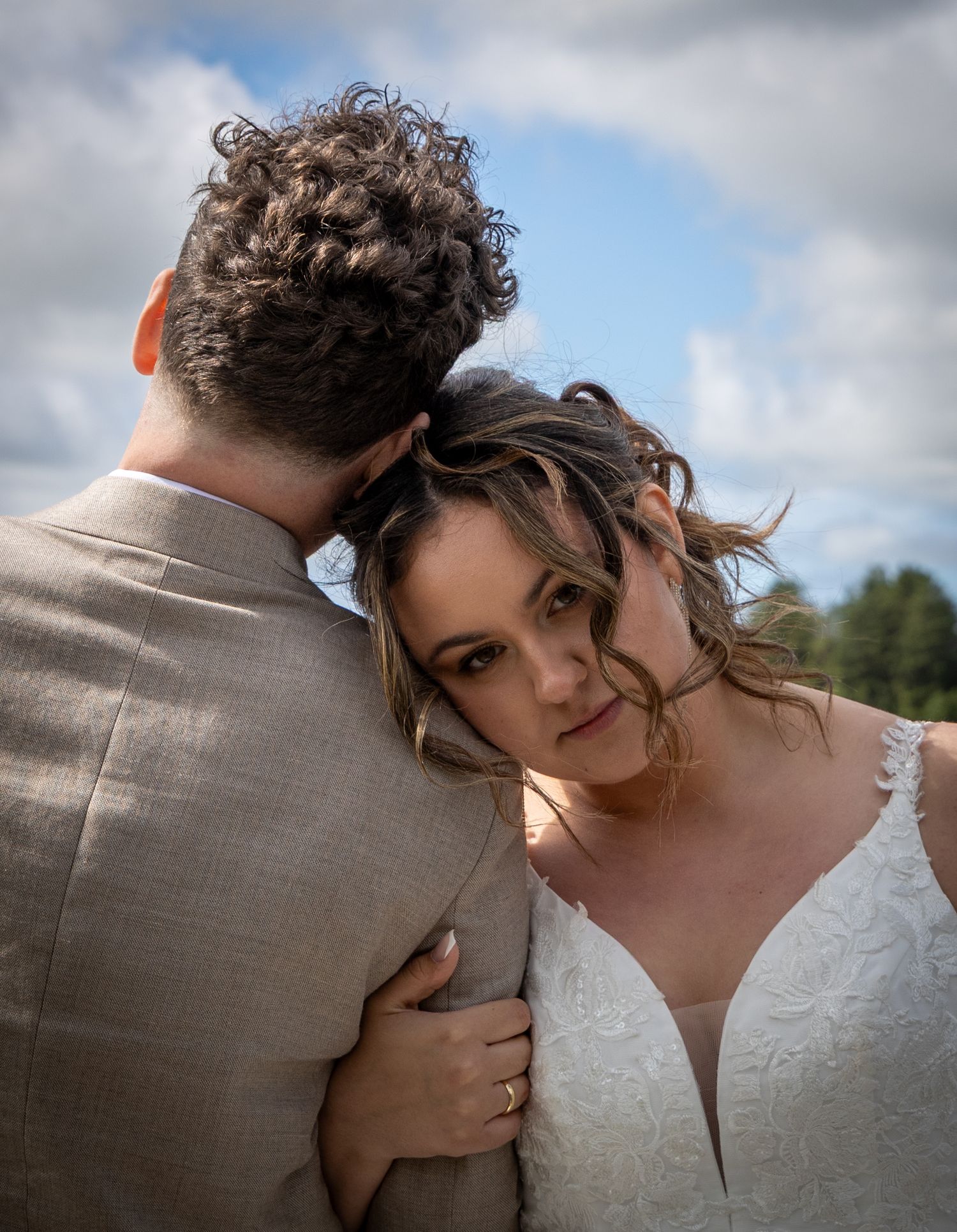 Bride in lace wedding gown rests her head on groom's shoulder under a cloudy sky.