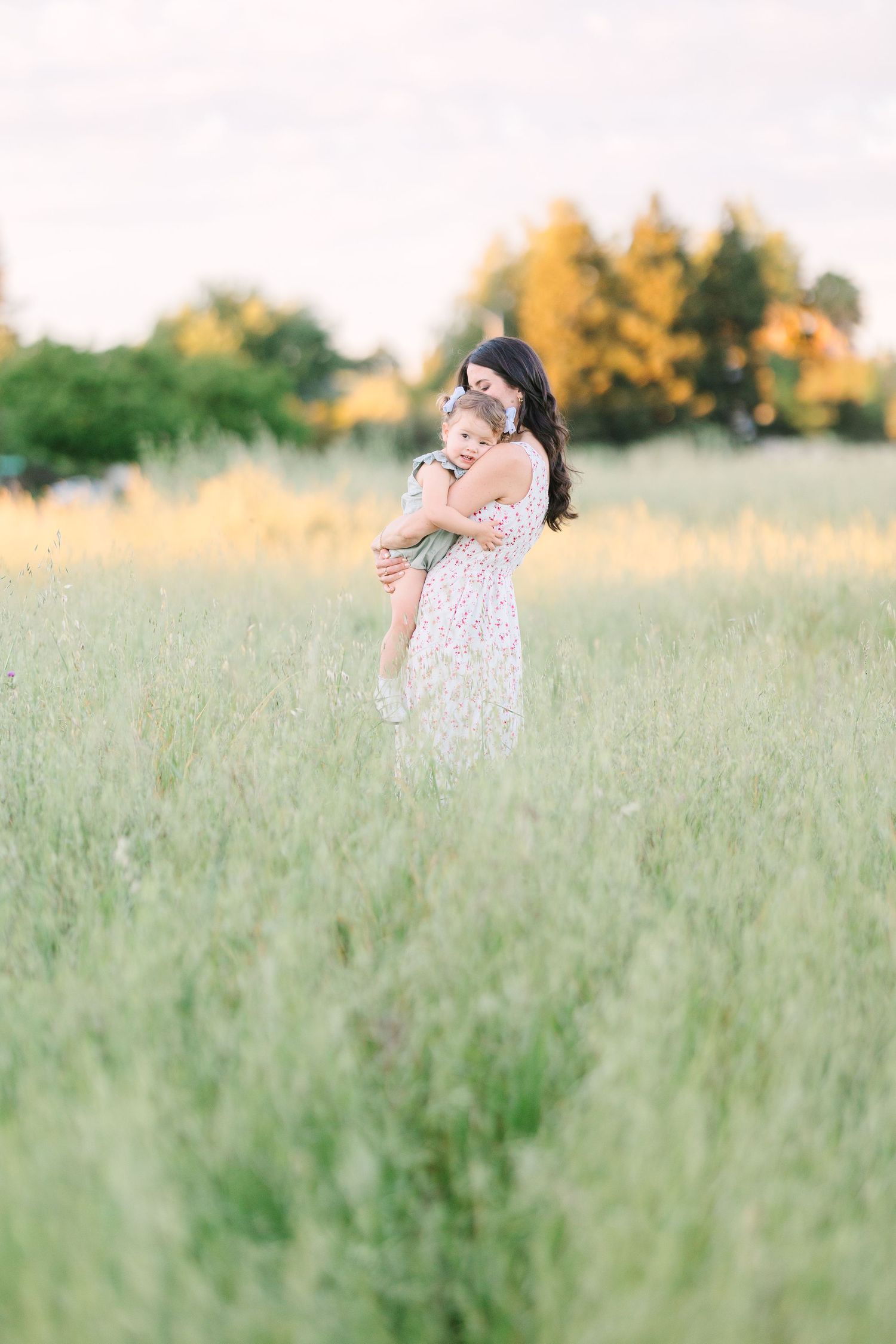 A mother in a floral dress holds her child in a sunlit green field with trees in the background.