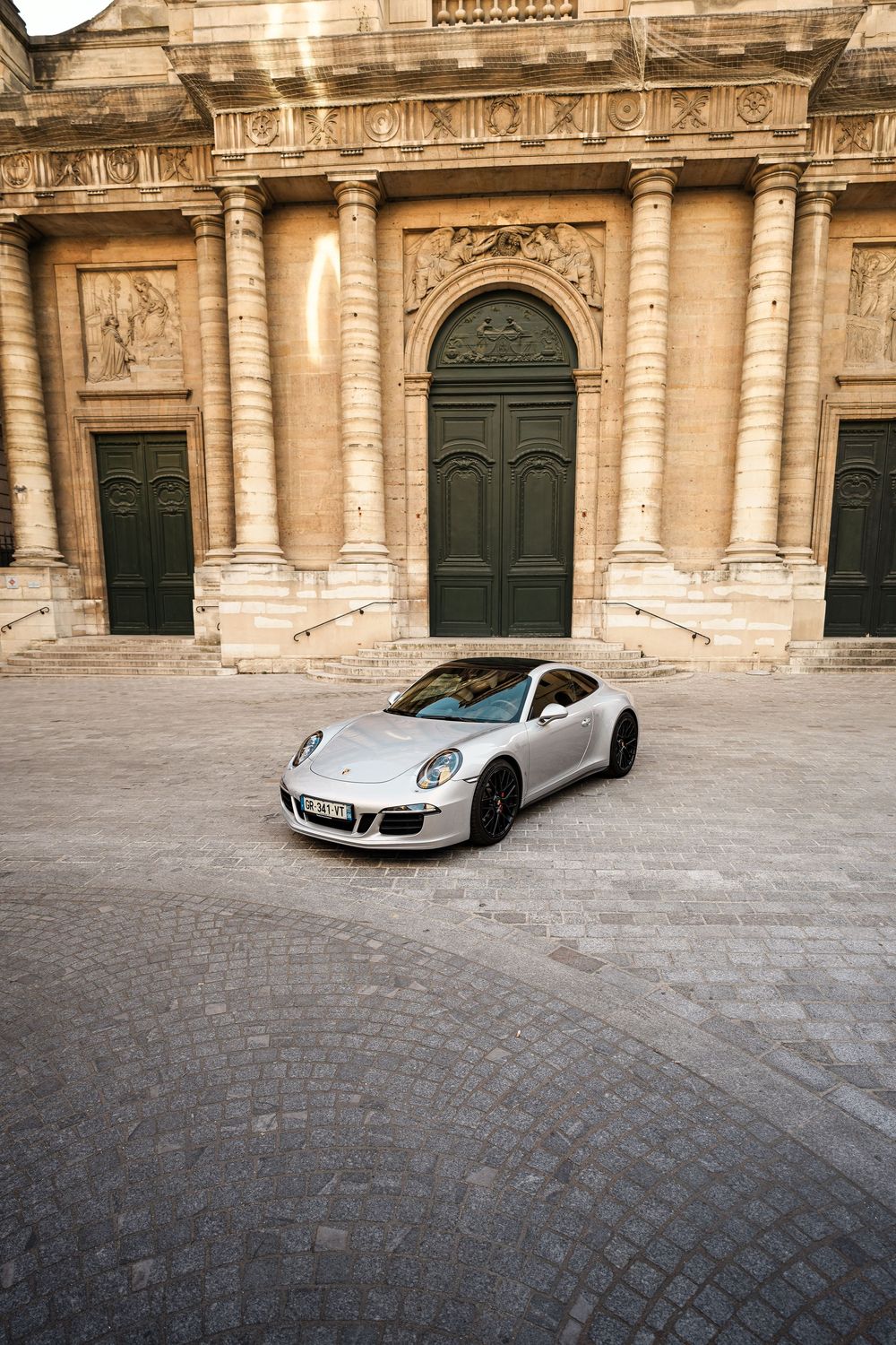 Silver Porsche 911 parked on cobblestones in front of a grand classical stone building with arched doors.