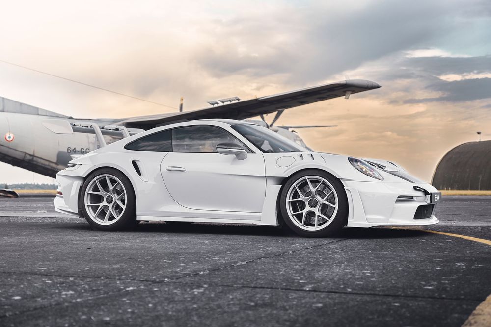 White Porsche 911 GT3 parked on tarmac near aircraft hangar with dramatic cloudy sky backdrop.