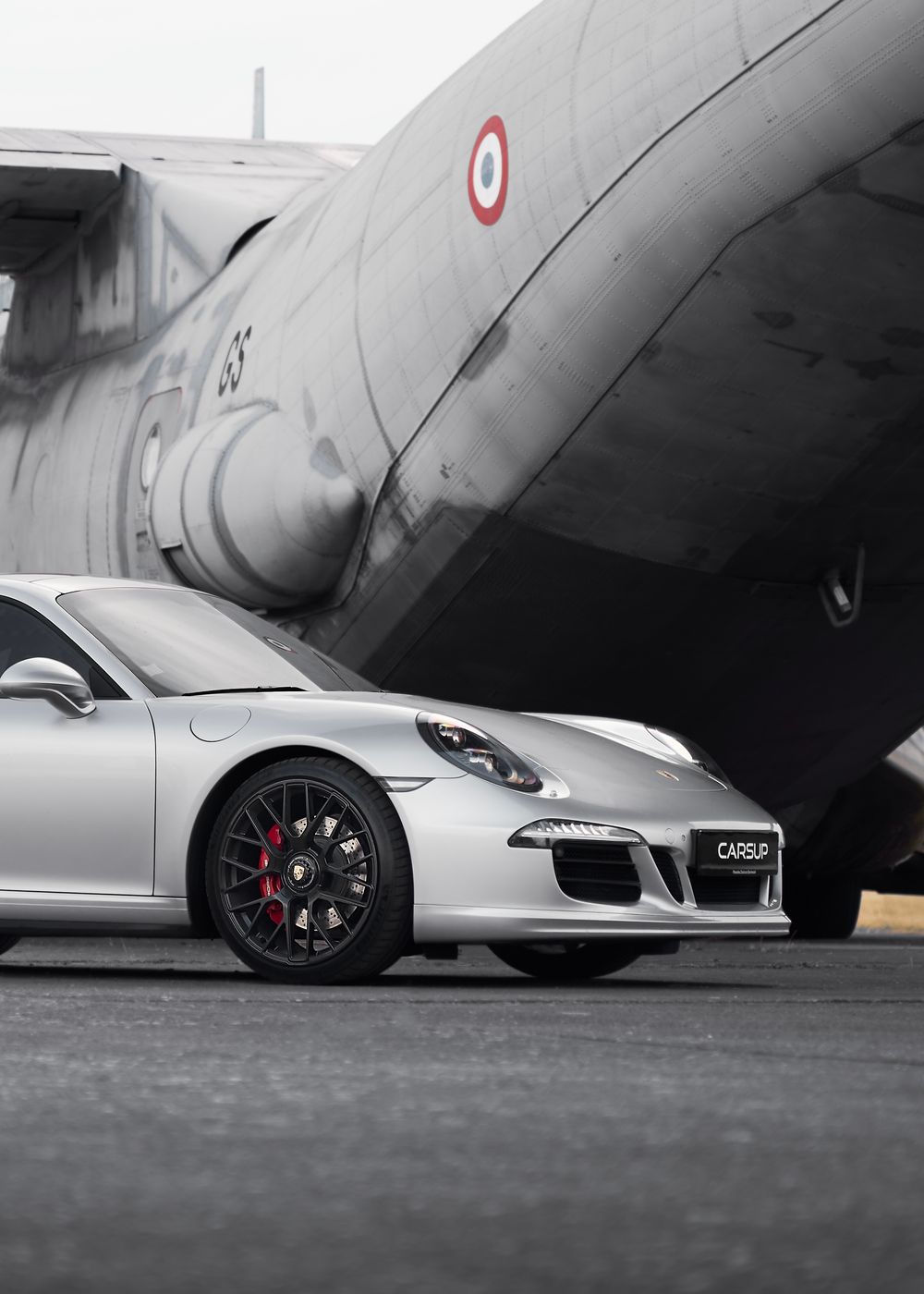 Silver Porsche 911 GTS parked beside a vintage military aircraft on a tarmac, showcasing luxury sports car design.