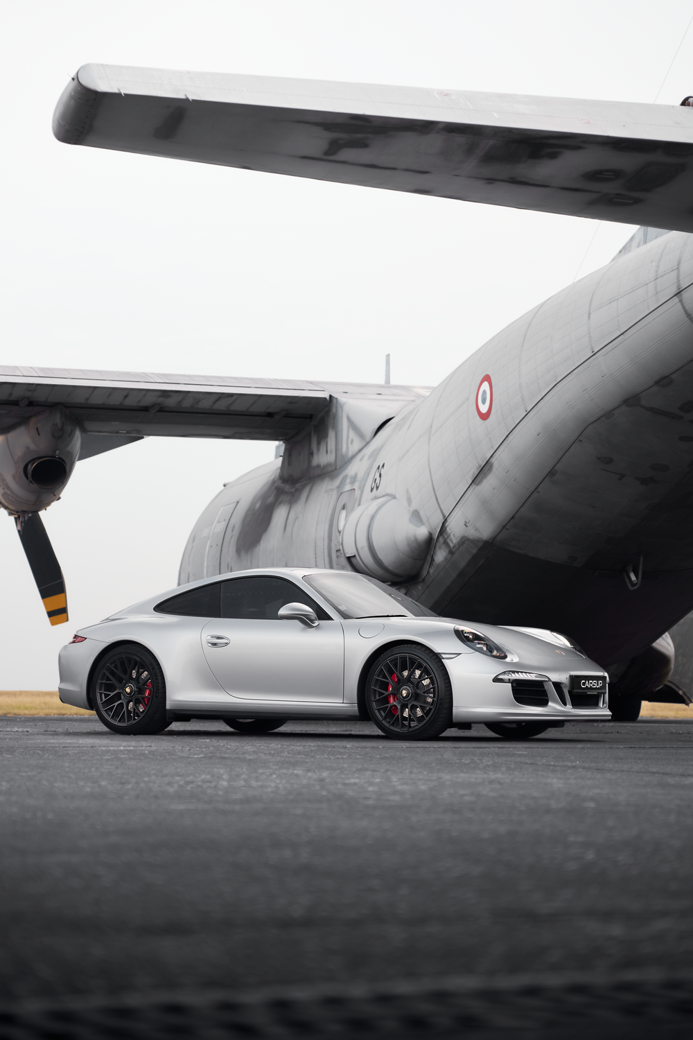 Silver Porsche 911 Carrera parked on tarmac beside a large military aircraft, showcasing its sleek design.