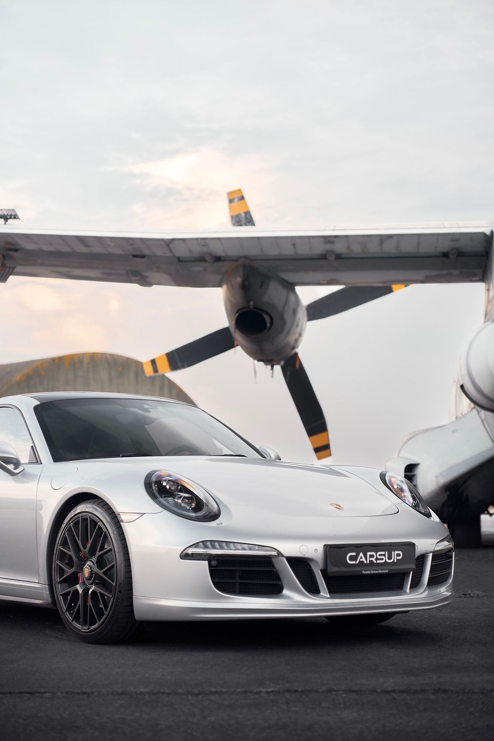 Silver Porsche 911 parked on an airfield tarmac with a vintage propeller aircraft in the background.