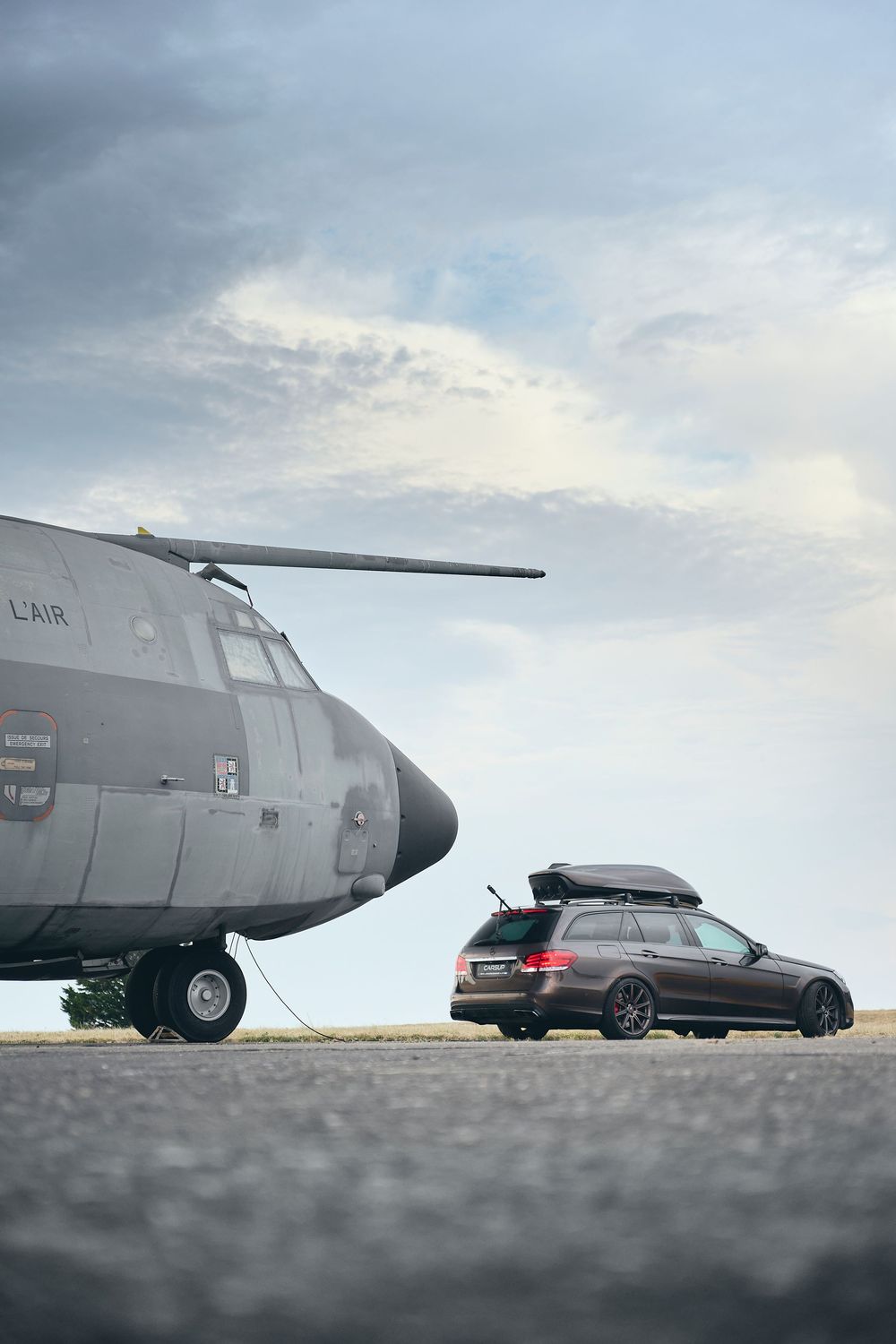 A military transport aircraft parked on a tarmac beside a dark estate car with a roof box under cloudy skies.