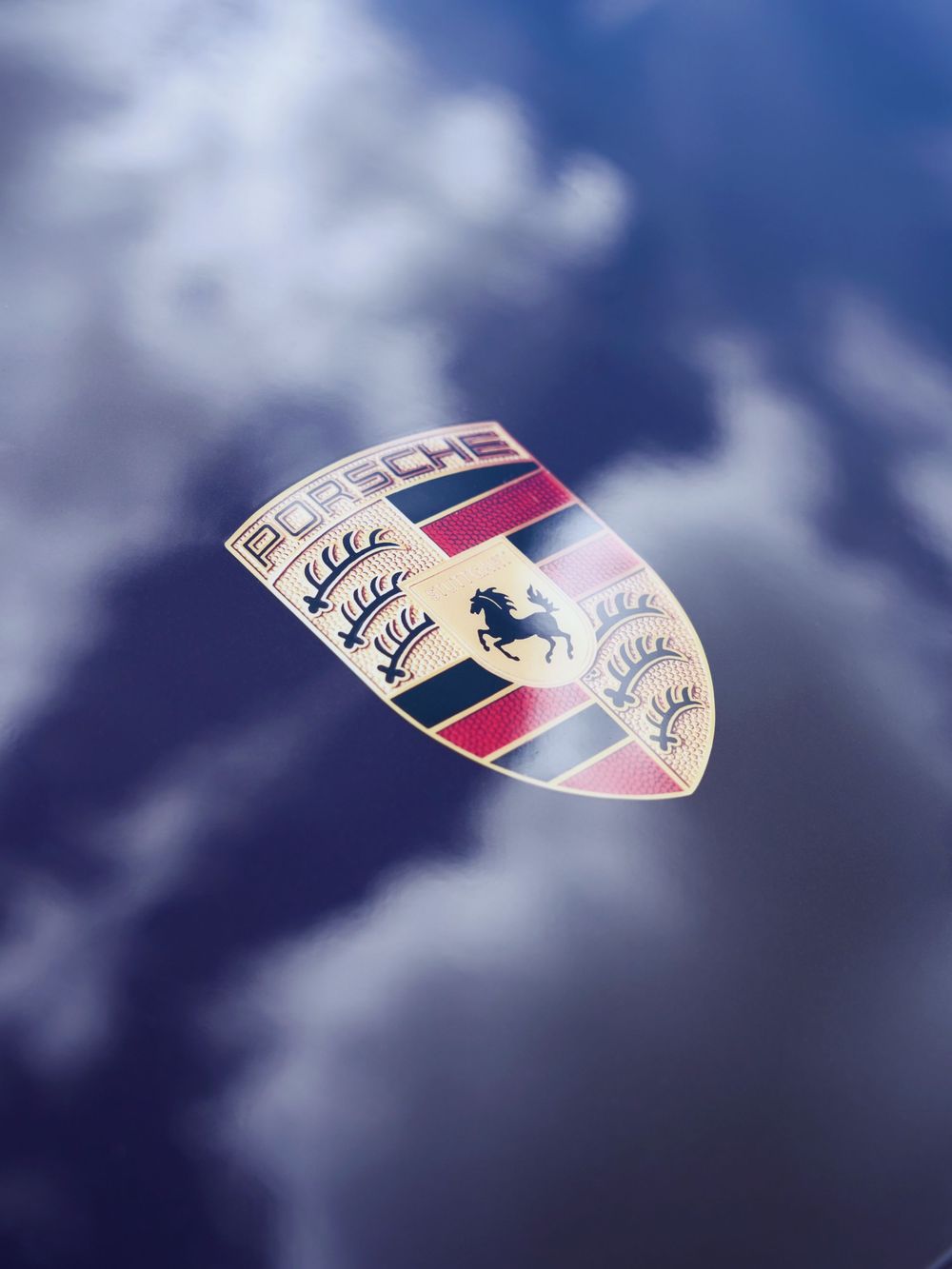 Close-up of the iconic Porsche crest badge reflecting cloudy blue skies, featuring a rearing horse and crown emblems.