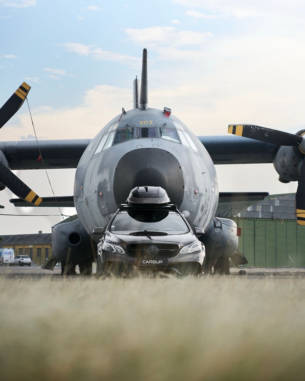 A sleek car parked in front of a large military propeller aircraft on an airfield runway.