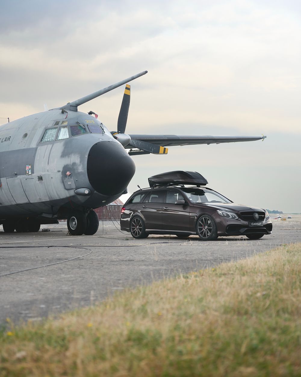 Dark Mercedes-Benz wagon with roof box parked on tarmac beside a military transport aircraft under cloudy skies.