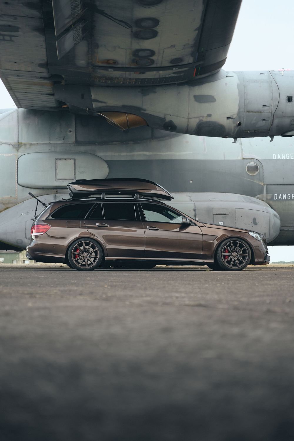 Brown Mercedes-Benz E-Class wagon with roof box and red brake calipers parked beside a military aircraft on tarmac.