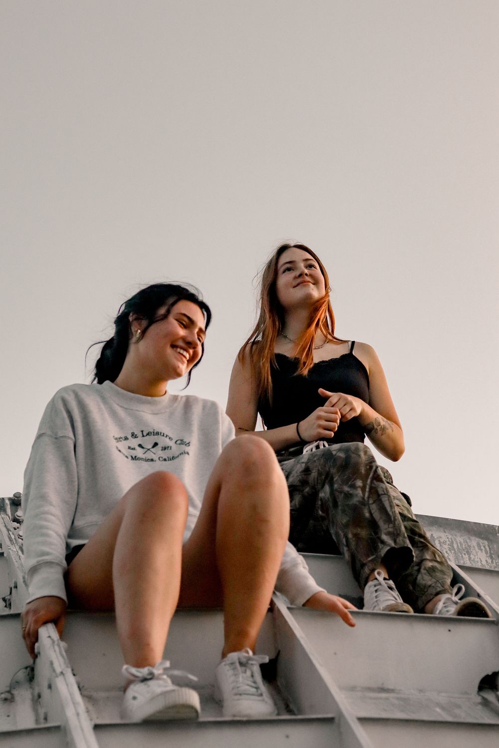 Two young women sitting outdoors on bleachers, smiling and laughing together at sunset.