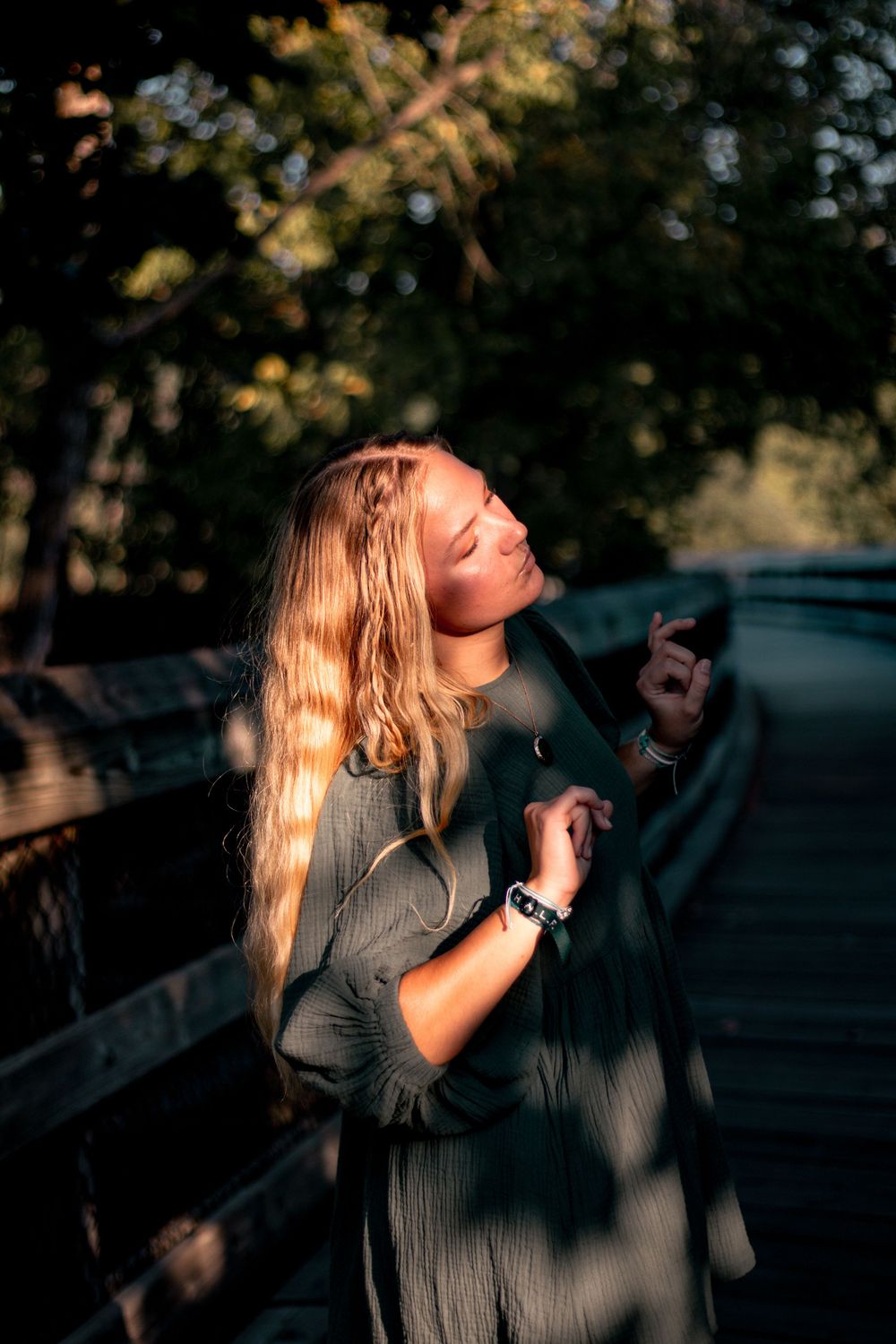 Young blonde woman standing on a wooden bridge in golden hour sunlight, eyes closed, wearing a gray dress.