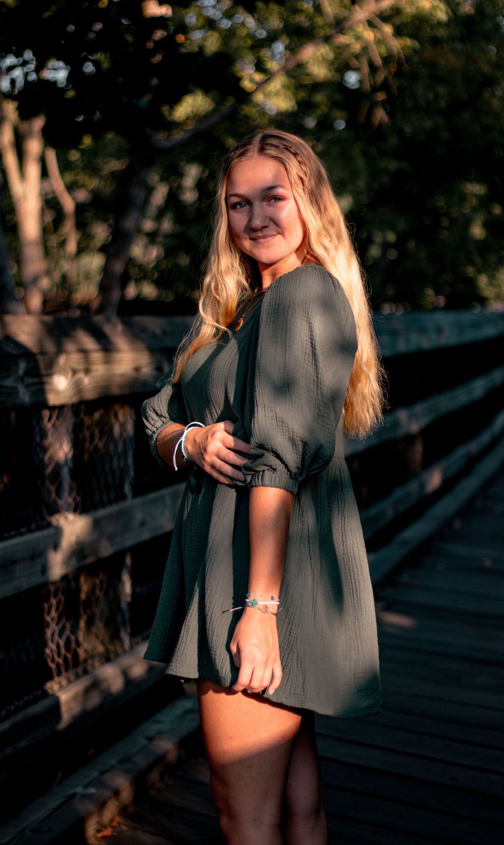 Young blonde woman in green mini dress posing on wooden bridge with sunlit trees in background.