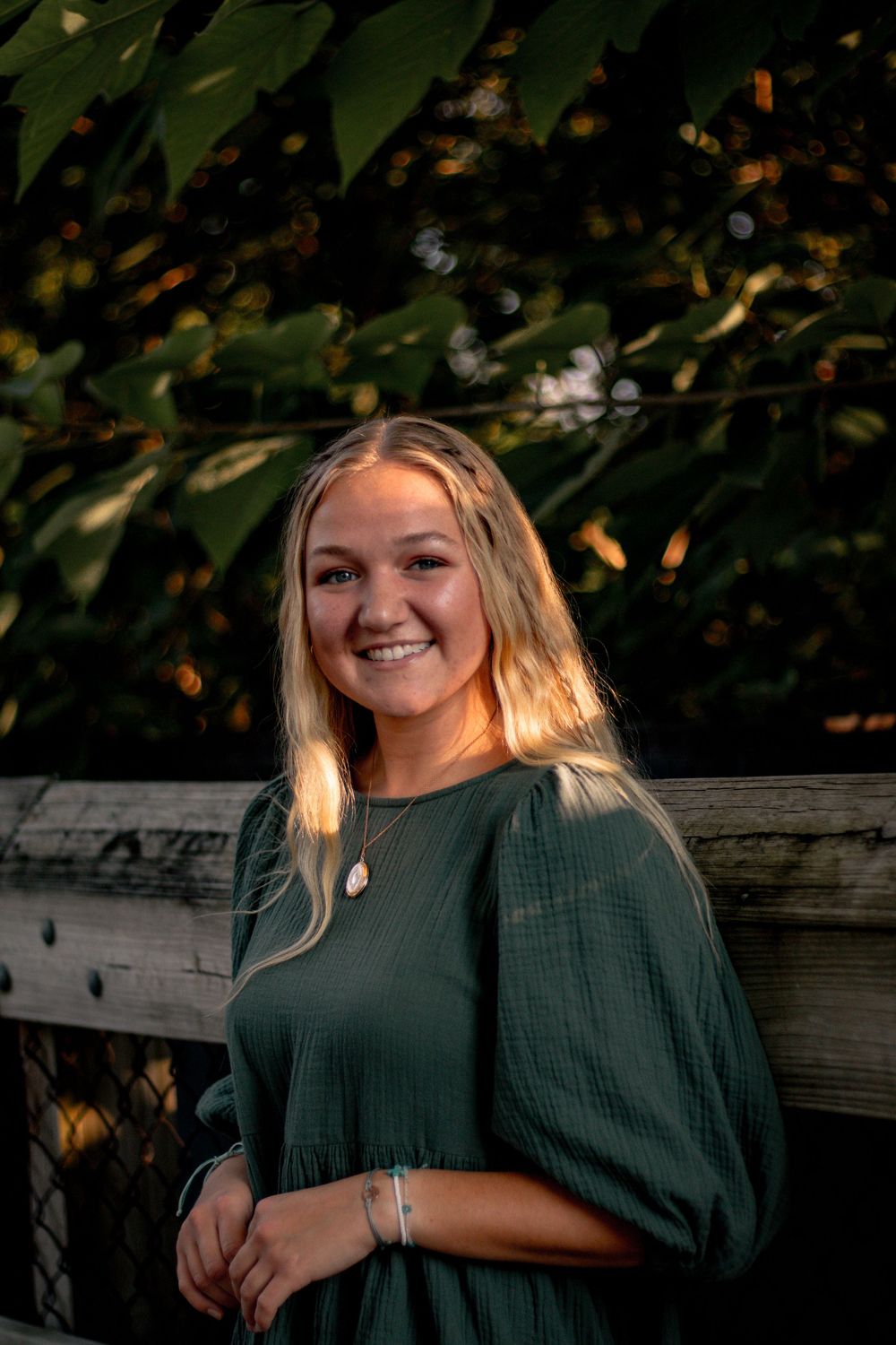 Young blonde woman smiling in green dress outdoors, golden hour light with lush green foliage background.