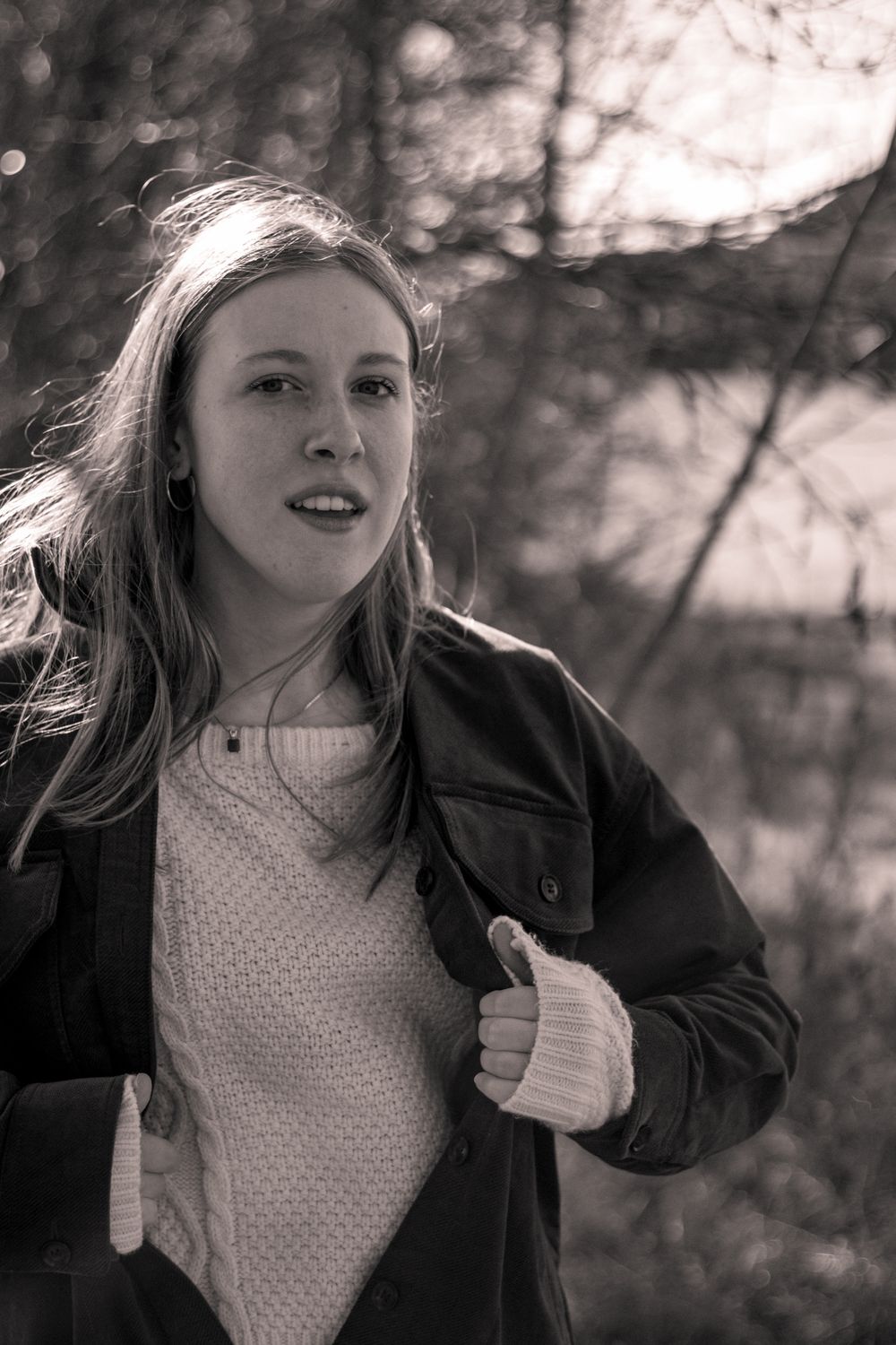 Black and white portrait of a young woman with long hair wearing a jacket outdoors among trees.