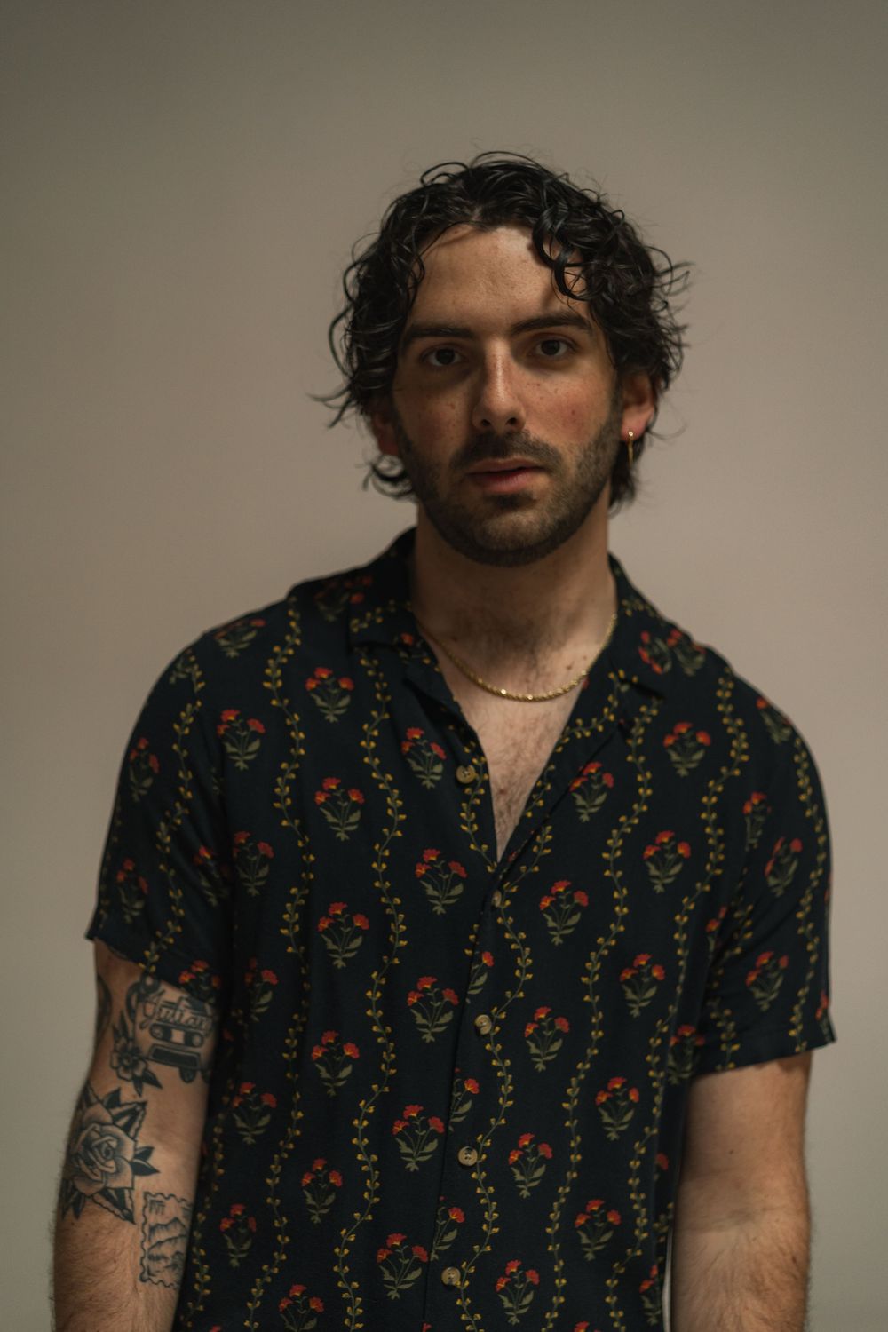 Young man with curly dark hair, tattoos, and floral print shirt posing against a neutral gray background.