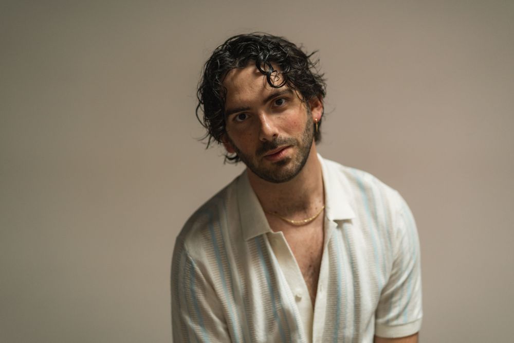 Young man with dark curly hair wearing a white button-up shirt and gold necklace posing against a gray background.