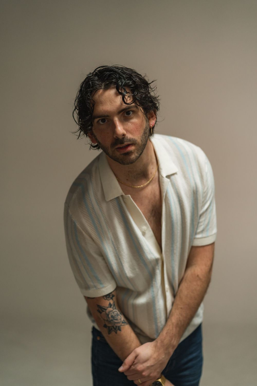 Young man with dark curly hair and arm tattoo poses in white striped polo shirt against neutral gray background.