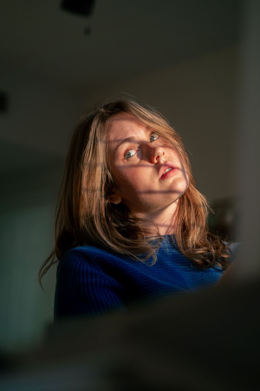 Young woman with long brown hair in blue sweater gazes upward, dramatically lit by natural light against dark background.
