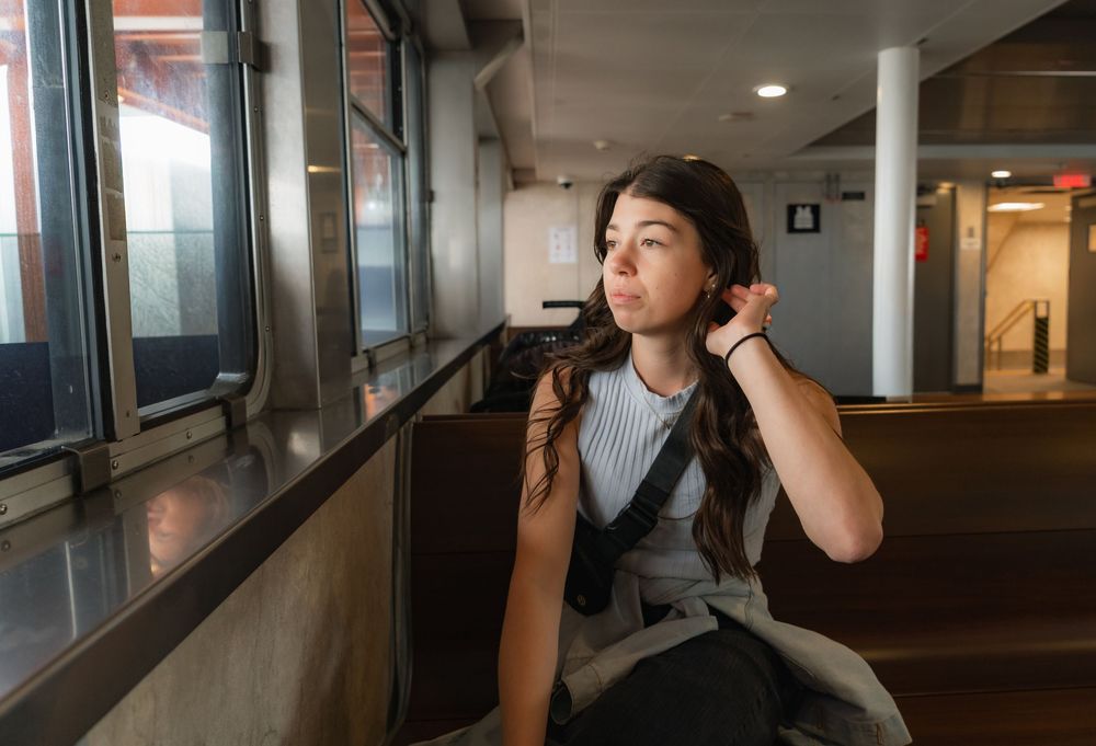 Young woman with dark hair sits by window in modern building, touching her hair thoughtfully.