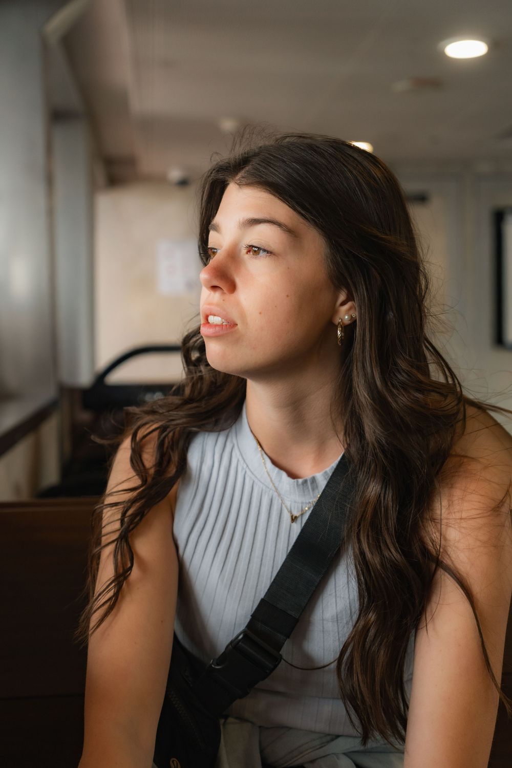 Young woman with long dark hair wearing a light blue sleeveless top and crossbody bag, gazing thoughtfully to the side.