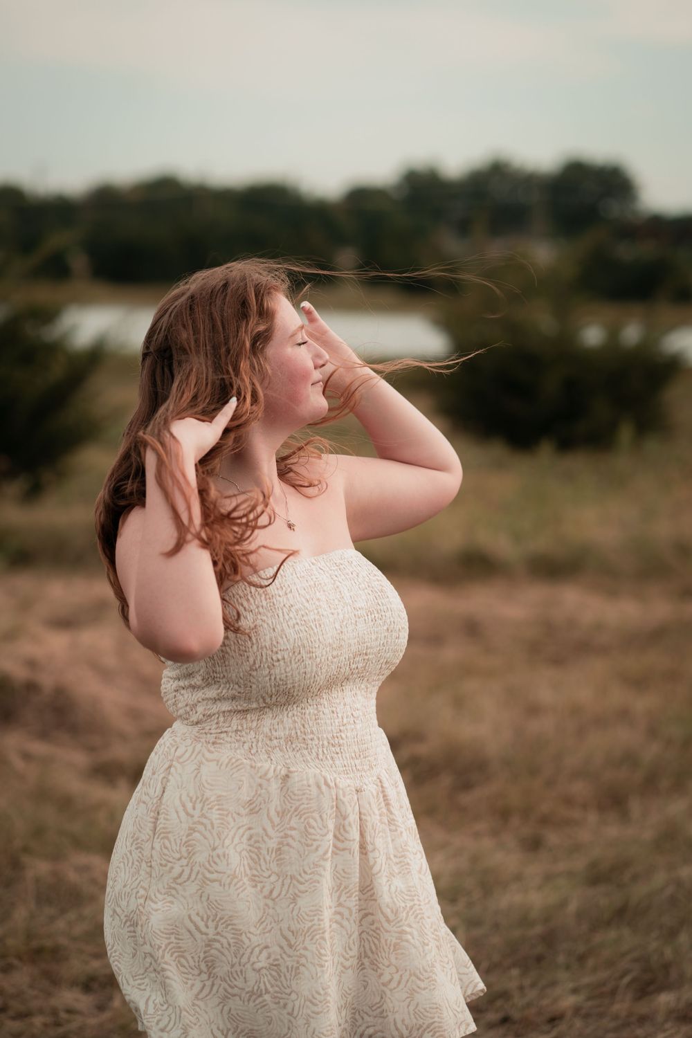 Young woman in white sundress standing in golden field, red hair blowing in wind, trees and lake in background.