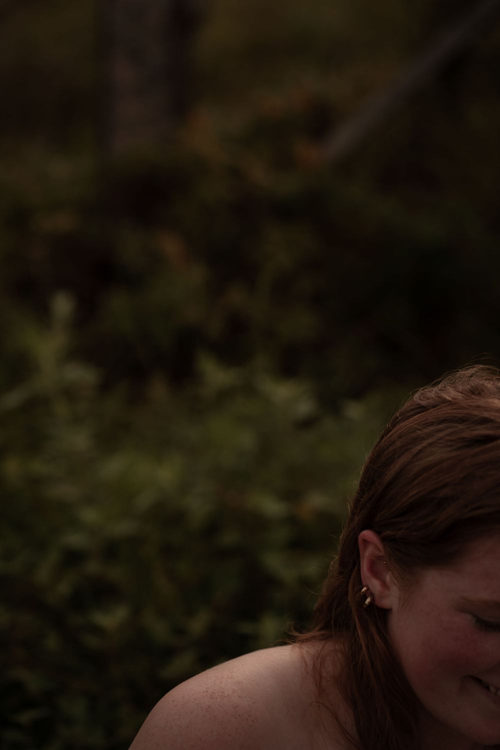 A young woman with red hair and small earrings looks down, set against a lush green forest background.