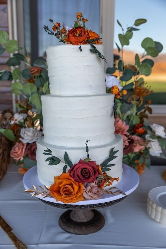 Three-tier white wedding cake with orange and red roses on wooden stand, surrounded by eucalyptus and floral arrangements.