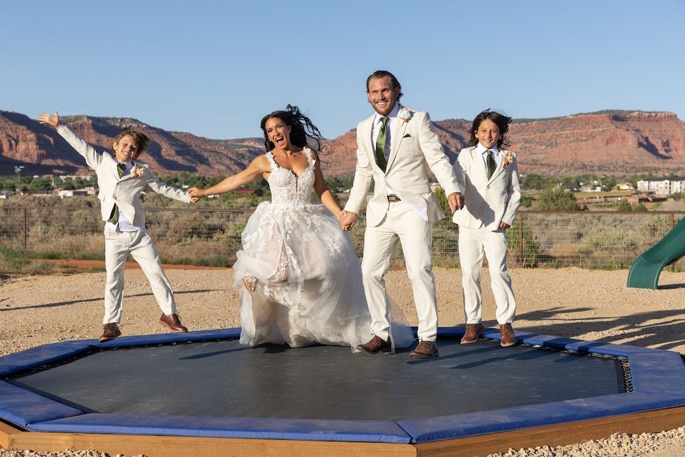 Bride and groom jumping on trampoline with two children in desert landscape during outdoor wedding celebration.