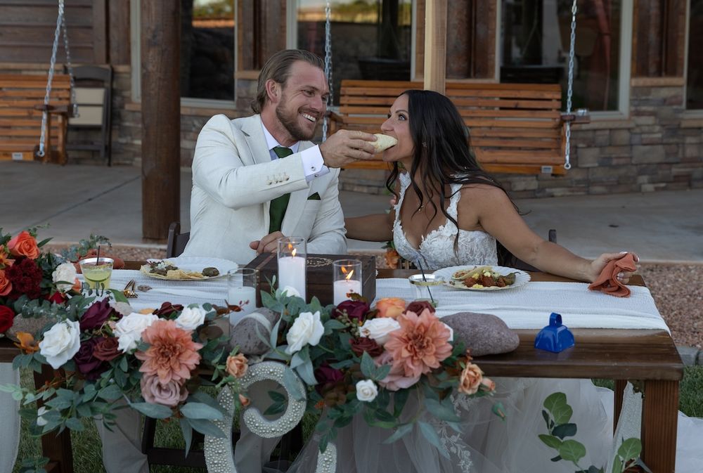 Bride and groom laughing at sweetheart table adorned with burgundy and blush floral arrangements at outdoor wedding reception.