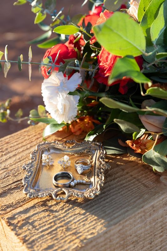 Wedding rings and earrings on ornate silver tray beside red and white floral bouquet on rustic wooden surface.
