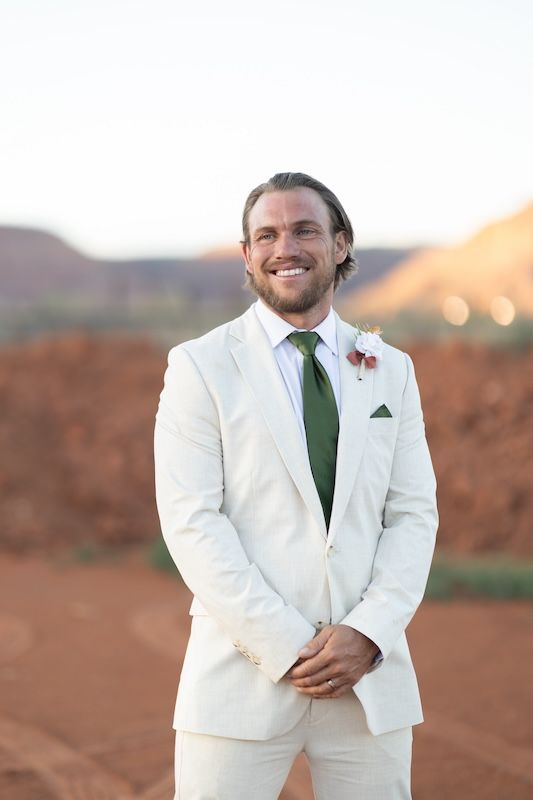 Smiling groom in white suit with green tie and boutonniere poses outdoors against red rock desert landscape.