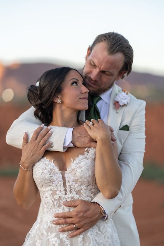Bride and groom embrace at sunset in red rock desert, wearing lace wedding gown and cream suit with boutonniere.