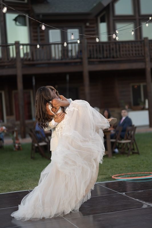 Groom lifting bride in white tulle gown during outdoor wedding first dance under string lights at rustic venue.