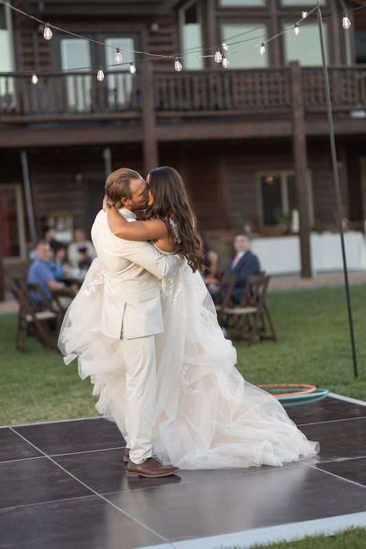 Bride and groom share first dance kiss outdoors on black dance floor at rustic wedding reception with string lights.