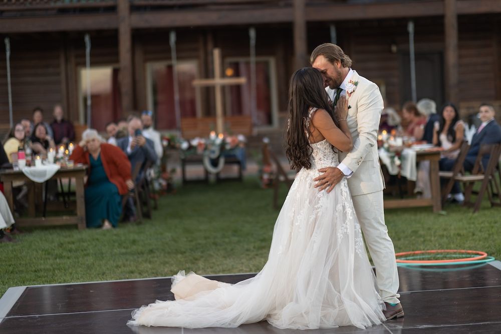 Bride and groom share first dance on outdoor floor at rustic wedding reception with guests seated at tables.