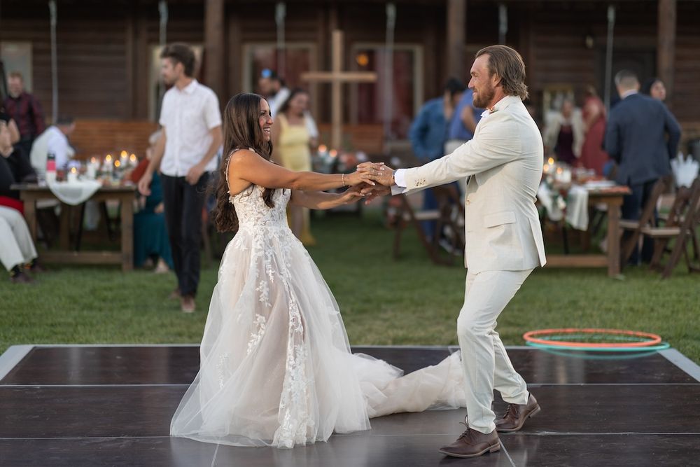 Bride and groom share first dance on outdoor stage at rustic wedding reception with guests dining in background.